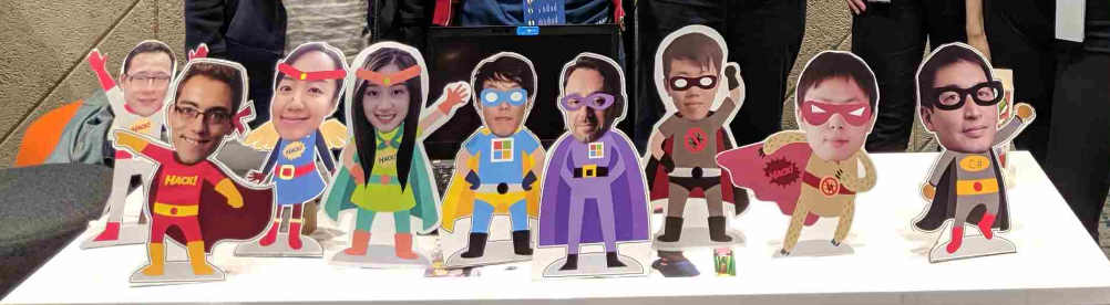Small cardboard cutouts of VROOM hackathon team members propped up on a table. Each cutout has a photograph of the team member’s face paired with a cartoon superhero body.