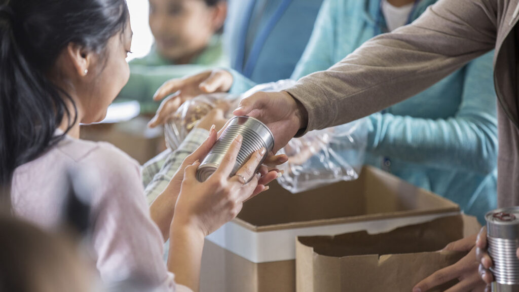 societal individual resilience image: Young woman receives a canned food item from food bank. An unrecognizable volunteer hands her the canned food item.