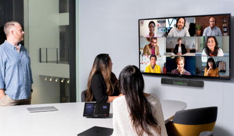 One male and two females in medium conference room with a Crestron Microsoft Teams Rooms on Android device and touch display being used for remote video meeting with Teams Meetings. Large mounted display showing remote participants and two Surface Device