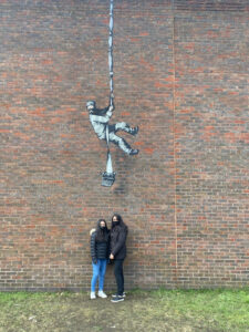 two people stood in front of the artwork. The artwork is high up on the wall, over head height. The wall is about four times as tall as the people.