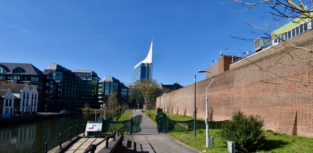 view along Oscar Wilde Memorial Walk. Along the right hand side is the tall wall of the prison, on the left is a canal. In the distance there are some office buildings and one tall, modern, spire shaped building which stands above all the others