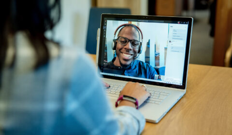 Woman at a desk using a Surface laptop to make a Microsoft Teams video call with one man smiling and wearing a headset. Business Voice conference call/meeting device is in the background.