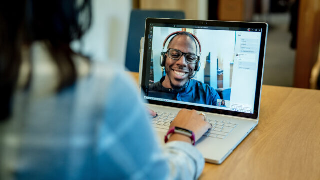 Woman at a desk using a Surface laptop to make a Microsoft Teams video call with one man smiling and wearing a headset. Business Voice conference call/meeting device is in the background.