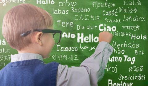 Photo of a young child writing on a chalkboard. The word “hello” is written on the chalkboard in multiple languages.