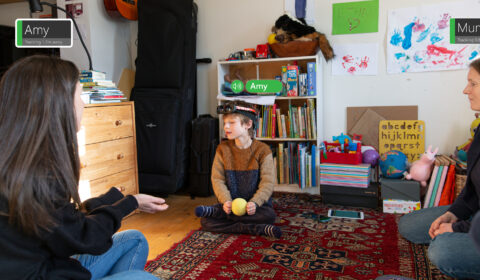 A young boy wearing the PeopleLens sits on the floor of a playroom holding a blind tennis ball in his hands. His attention is directed toward a woman sitting on the floor in front of him holding her hands out. The PeopleLens looks like small goggles that sit on the forehead. The image is marked with visual annotations to indicate what the PeopleLens is seeing and what sounds are being heard.