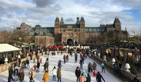 Photo of the Rijksmuseum, a huge grand building with a combination of gothic and renaissance architecture. In front of the museum are large letters spelling out ‘I Amsterdam’, surrounded by people ice skating.