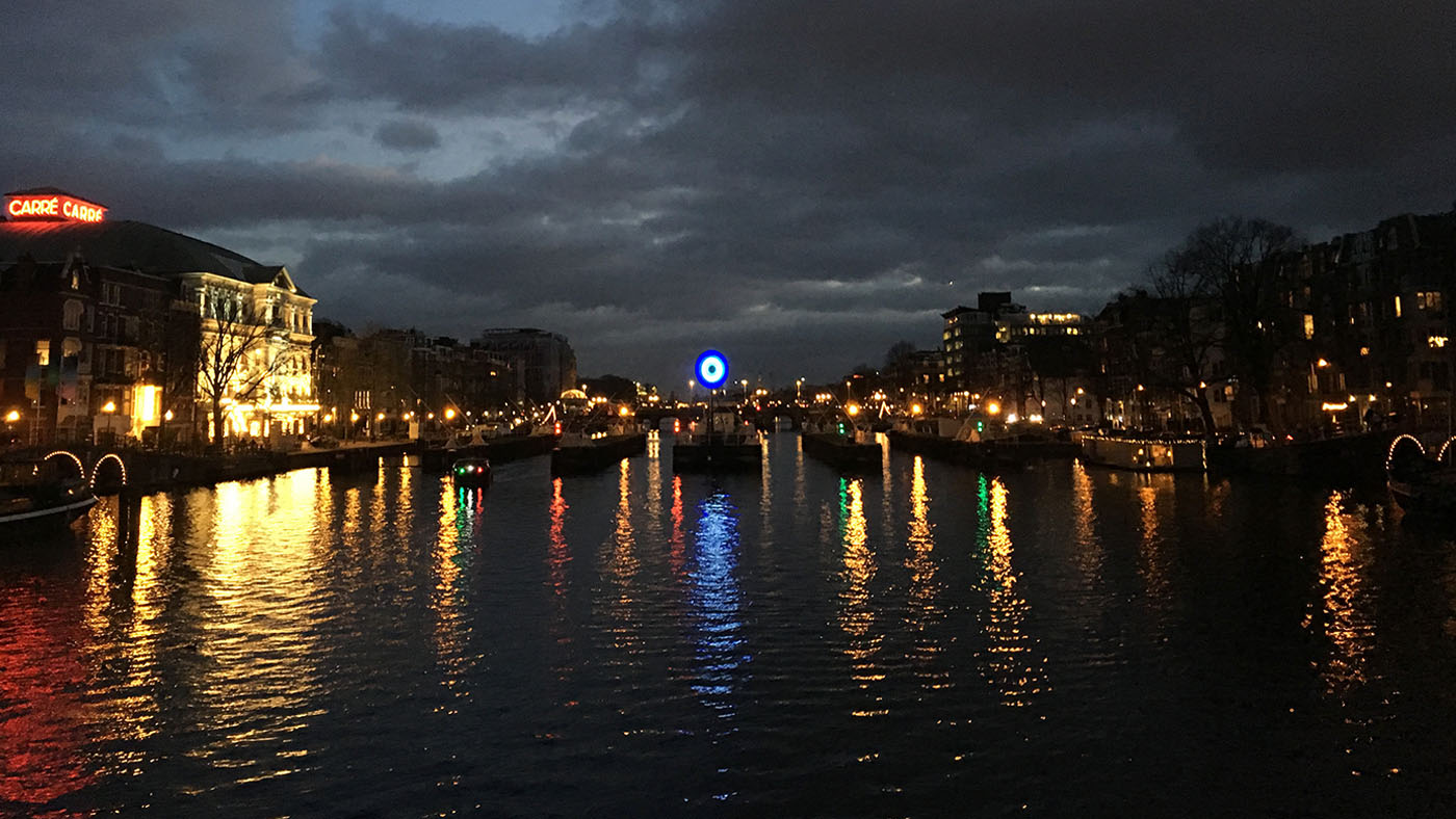 Soundscape - A view along a wide Amsterdam canal to a bridge at night-time. The bridge, and a building to the l eft of it, are both illuminated with the water reflecting off the surface of the water