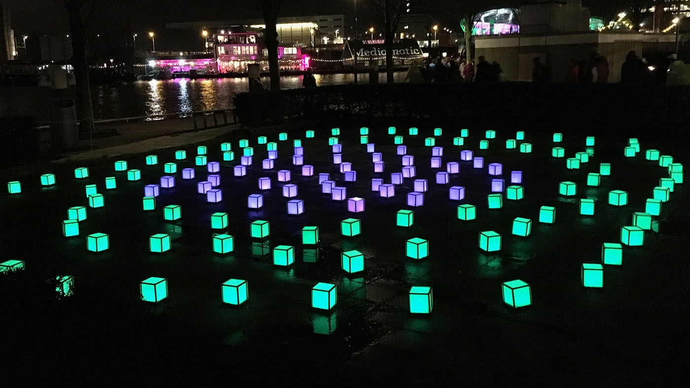 Soundscape - an image of a light installation in an Amsterdam canal. Several small, illuminated cube, arranged roughly in circles with the outer circles illuminated in green and the inner circles in purple.