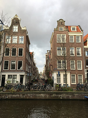 Soundscape - View across a canal and down a narrow. side street. There is a row of bikes parked on the opposite bank of the canal. The buildings are tall and narrow, 5 storeys high, with lots of windows.