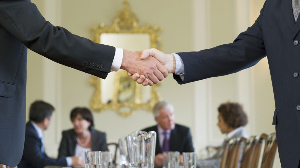 two men shaking hands over a table surrounded by other people talking amongst one another