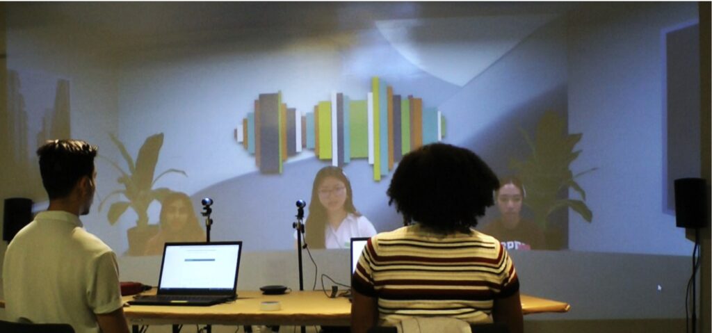 A hybrid Perspectives meeting with two local and three remote participants. The remote participants are projected onto a wall and look like they are part of the same virtual room connected to the local room.
