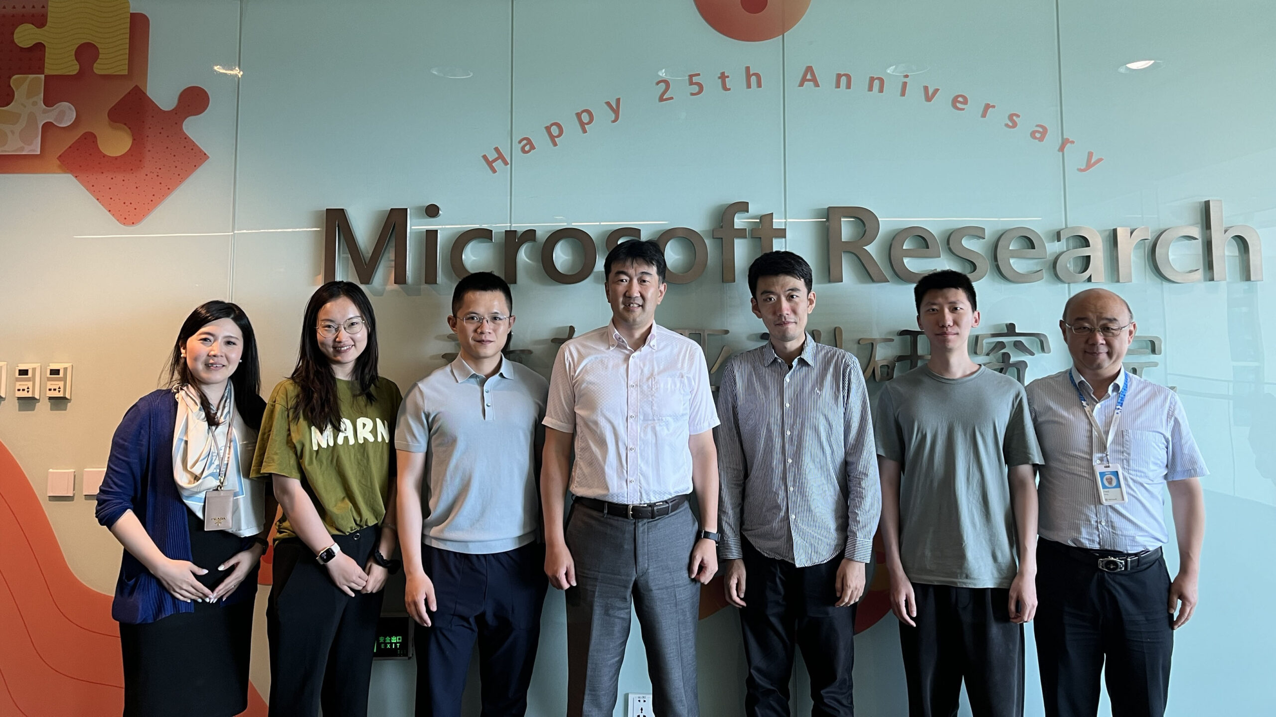 Seven people posed for a group photo in front of the wall banner of Microsoft Research Asia when Atsushi Ohma visited in June 2024.