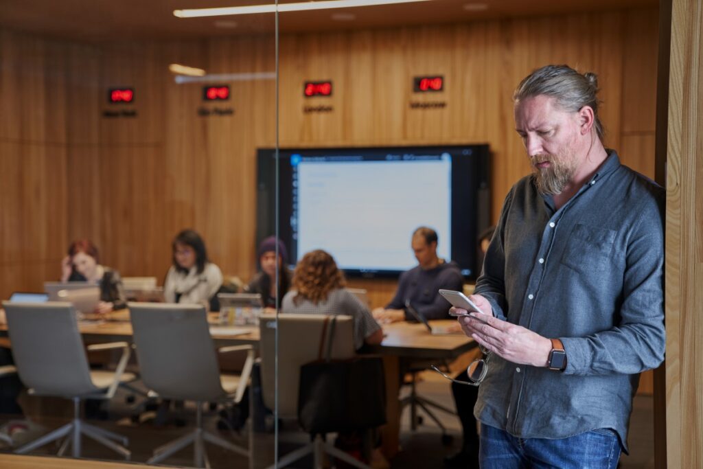 Bearded man looking at his phone with background of a blurry conference room of people working.