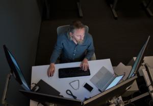 Top-down view of a bearded man in a gray/blue shirt seated at a desk working on a Surface laptop