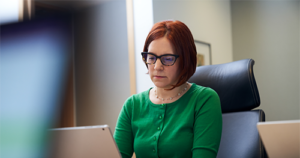 An image of a red-haired woman with a green shirt and glasses, sitting inside an office working on her laptop.