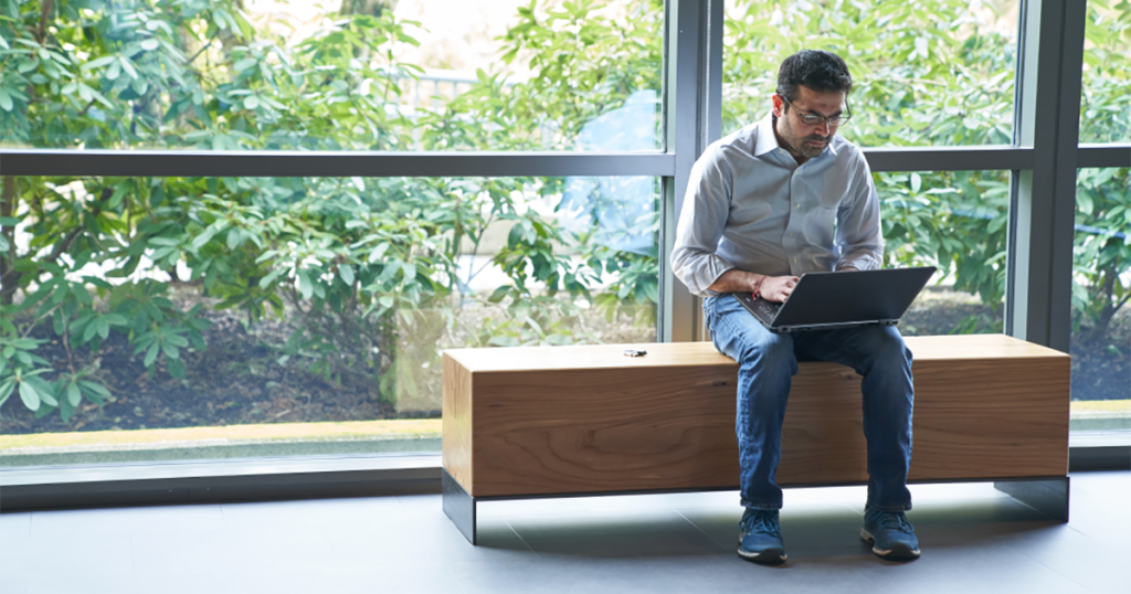 An image of a man sitting on an indoor bench in front of windows working on a laptop with a security key fob or USB drive next to him on the bench.
