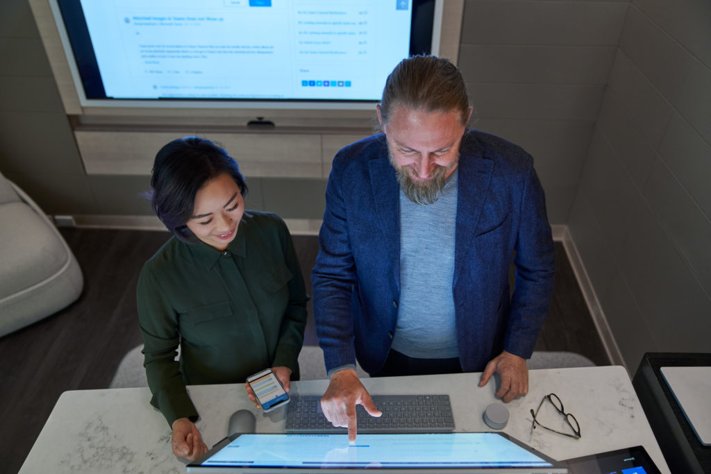 Top down view of a man and woman in a dim office collaborating or working on a Microsoft Surface Studio. She is holding a phone.