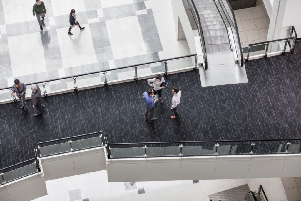 Aerial view of office building‘s interior entrance hall. Close-up of two separate groups of people standing together on an elevated walkway, chatting. Two others are walking on ground floor below.