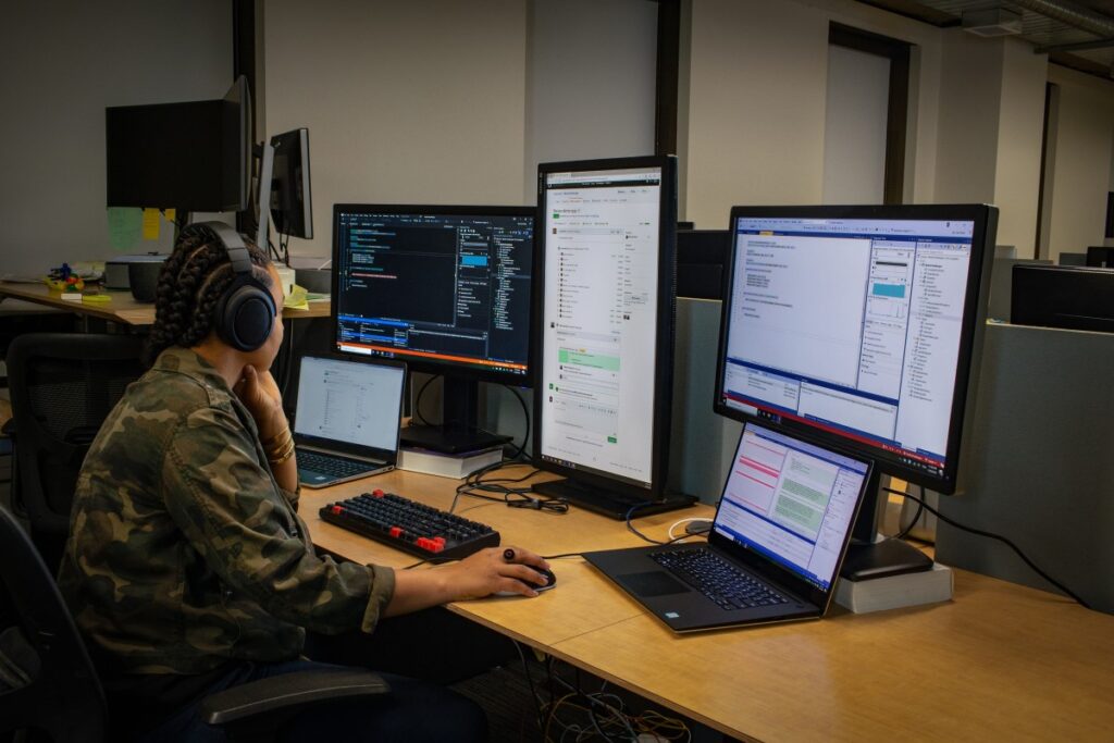 Black female developer wearing headphones; coding at her PC workspace in an enterprise office, using Visual Studio on a multi-monitor set up.