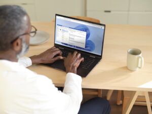 Man sitting at a wooden table typing on a laptop while using Microsoft Teams within Windows 365. Shown on Windows 11.