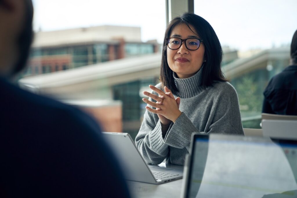 Woman with glasses sitting at a community table in common area looking up from behind a Surface laptop with her hands clasped together.