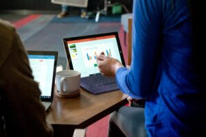 Two open laptops sitting on table in front of two figures, with a cup of coffee between them.