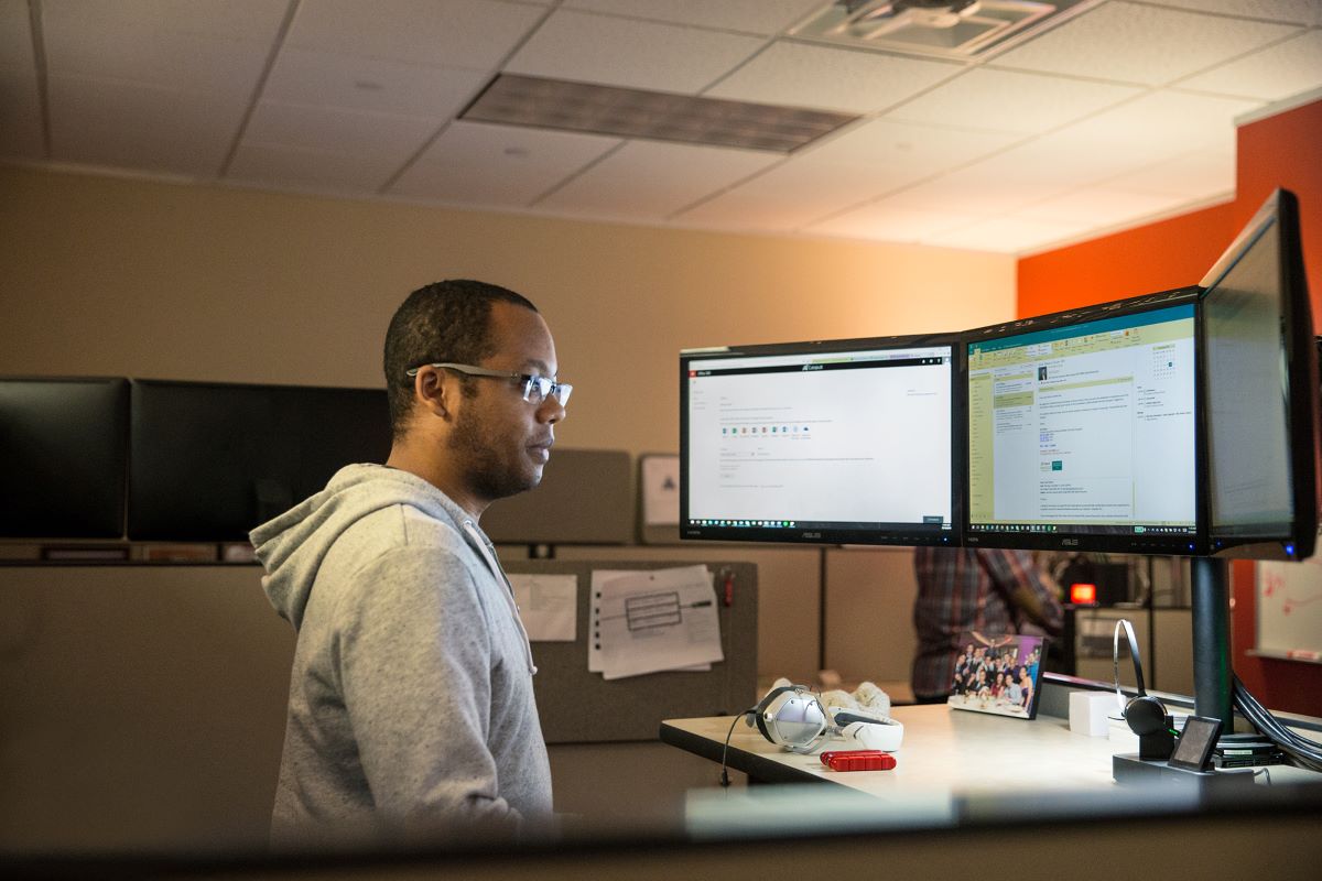 Photo of a male wearing a gray jacket and glasses, standing in front of a computer desk with multiple monitors.