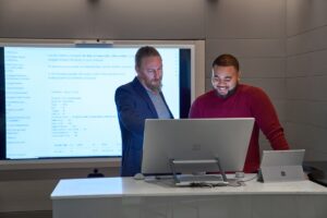 Forward facing view of two men working on a Microsoft Surface Studio with a larger blurred screen and display behind them.