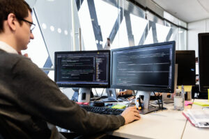 Male developer coding in front of two monitors at desk in office. Programming code shown on both monitors.