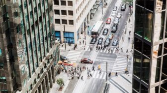 Downtown city street view from high angle inside office building. Large groups of pedestrians walk across street at crosswalk as car traffic flows through stoplight at busy intersection.