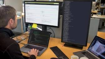 Photo of Male developer is coding at his workspace in an enterprise office using Visual Studio on a MacBook Pro multi-monitor set up.