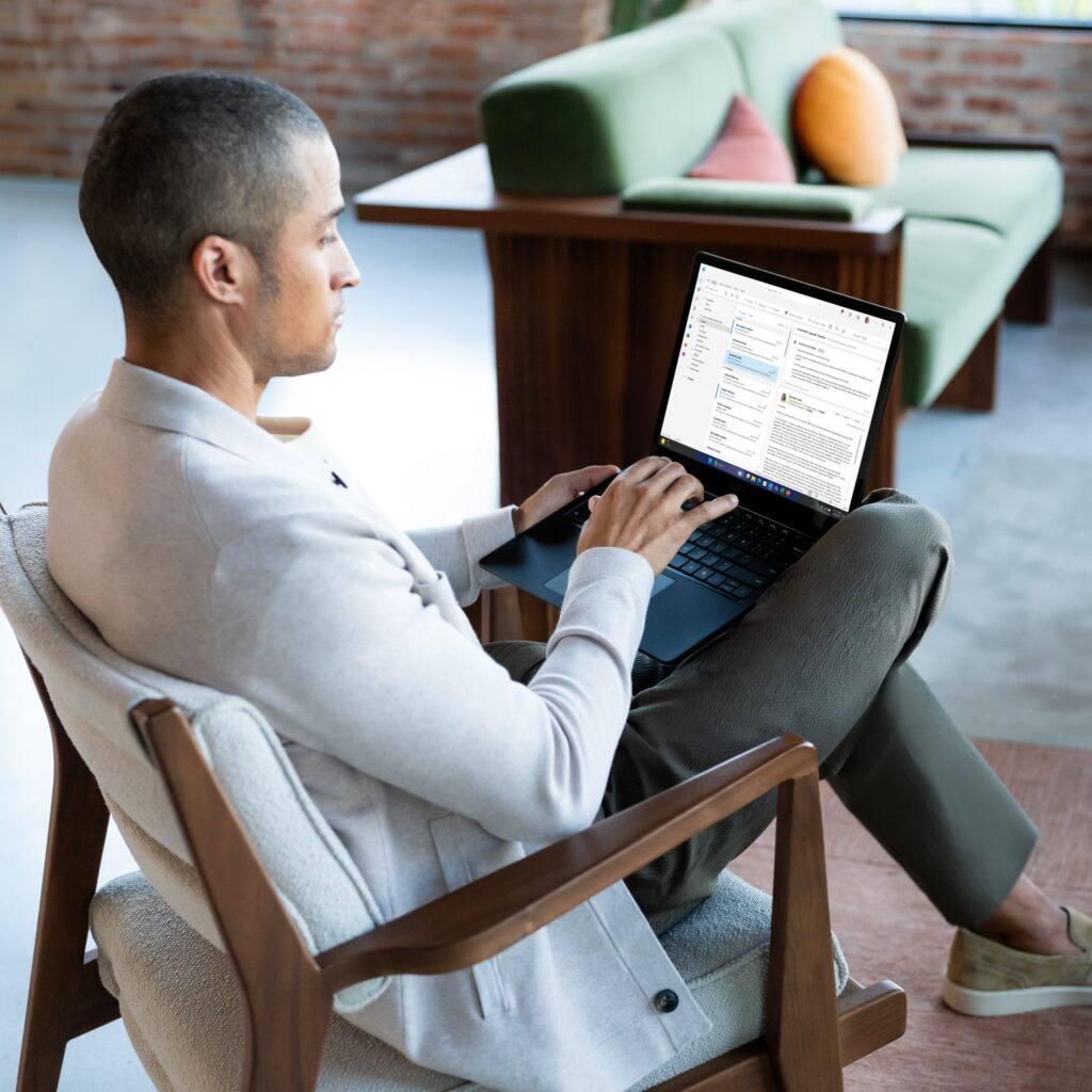 Photograph of male sitting on lobby chair collaborating on a Surface Laptop 6 in Black.