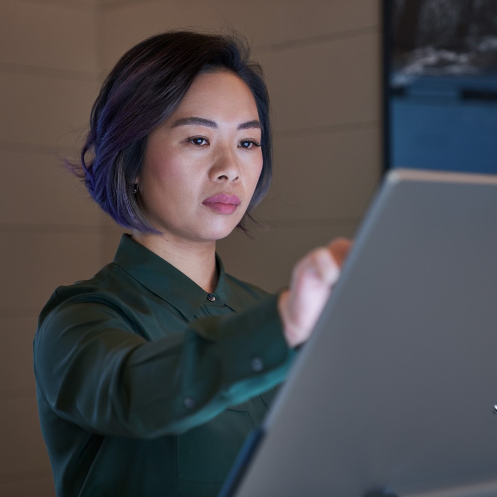 Side profile of a woman wearing a dark shirt in a dim office reaching up and working on a Microsoft Surface Studio.