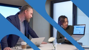 A woman and a man men working in a meeting room with Surface laptops. The woman is holding up her laptop and presenting her screen.