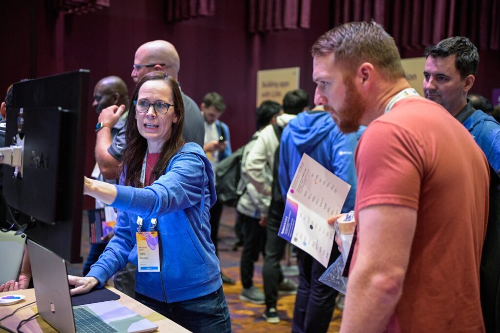 Two people at a conference look at a computer screen while a presenter points at it. 