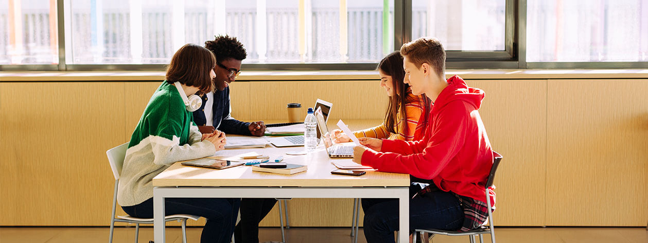 Keturi studentai sėdi prie stalo studijuoti ir kalbėti