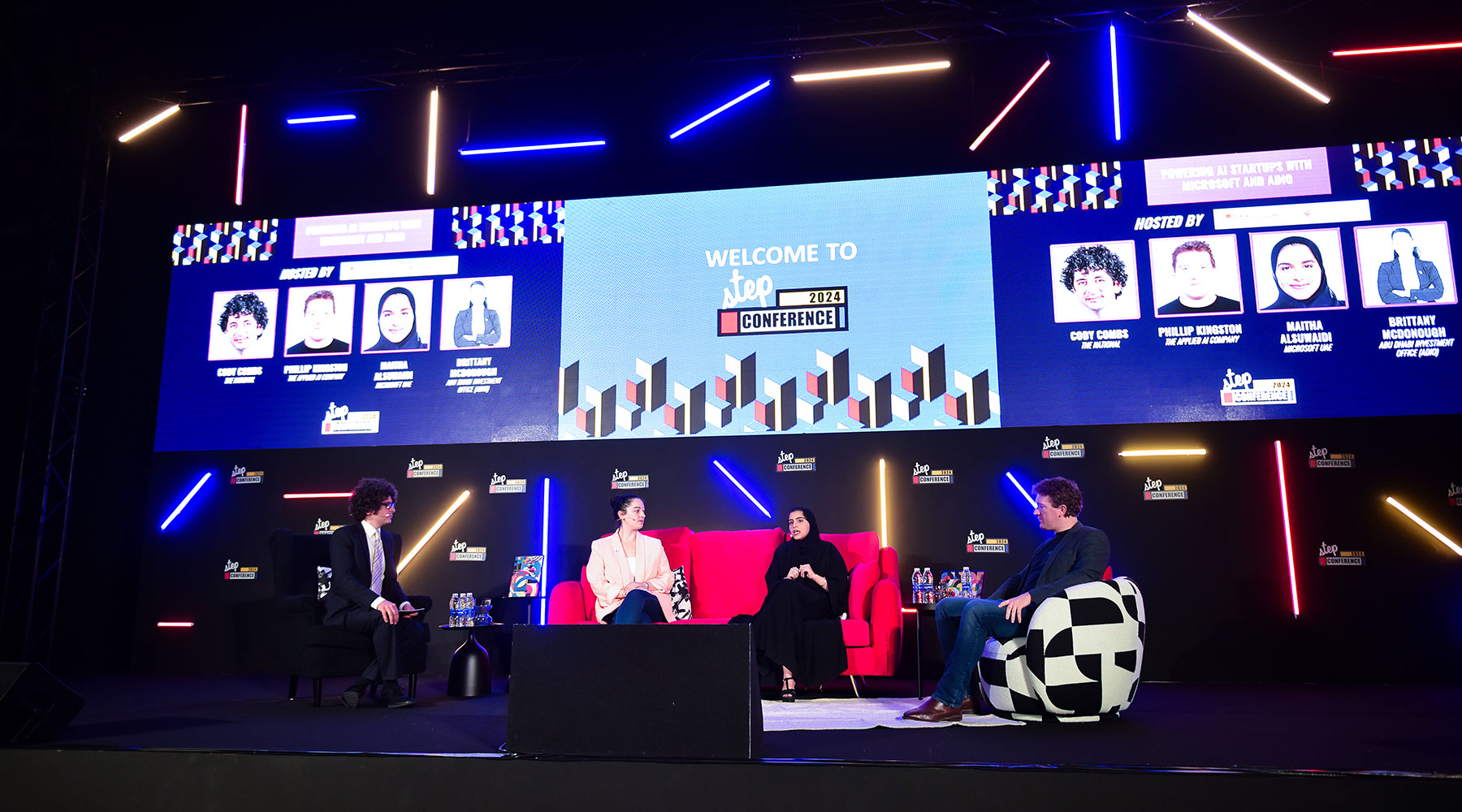 Group of people sitting down on a stage in chairs and talking with 3 screens in the background