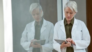 Mujer, de mediana edad, personal de salud sosteniendo una Tablet en sus manos. Se encuentra junto a un vidrio que sirve como espejo y la refleja.