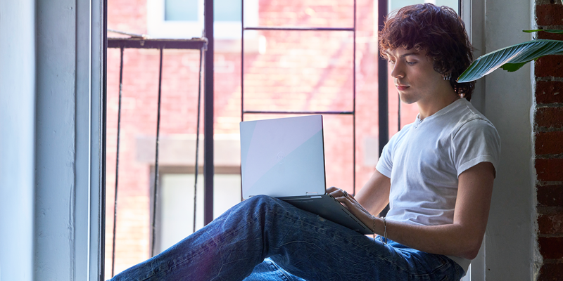 Kuvassa nuori mies istuu ikkunalaudalla sylissään kannettava tietokone