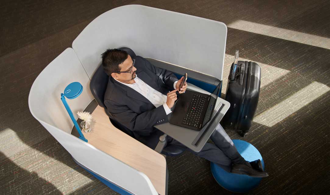 Top-down view of a man in a sport coat kicking his feet up and sitting in privacy table at the airport or hotel typing on his phone. A laptop is on the desk in the kiosk and a suitcase is near his feet