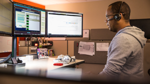 Image of a firstline worker speaking to a colleague over a headset while looking at several computer monitors.