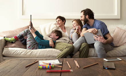 A family sits on a couch holding smart devices and laughing.