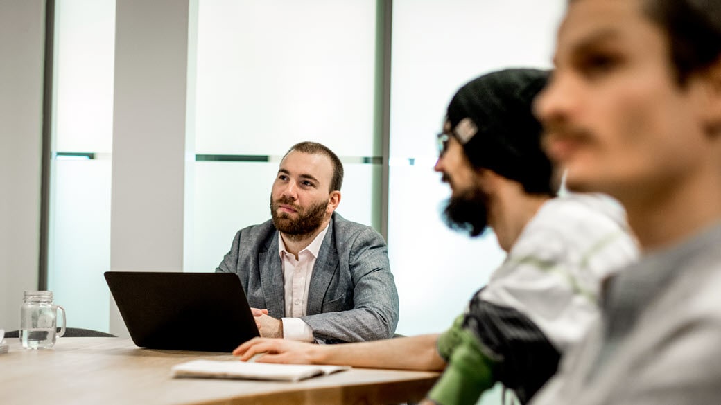 Three male employees sit at table in conference room of Ubisoft office. The employee at the head of the table has laptop in front of him.