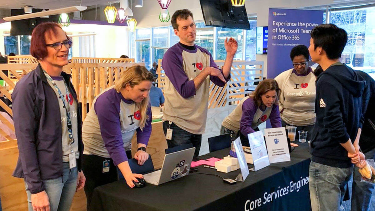 A photo illustrating a Teams Tuesday event. It shows members of the adoption team engaging with a Microsoft employee at a Teams Tuesday kiosk.