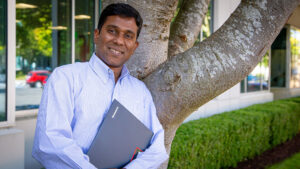 Raja Narayan looks at the camera with his hands holding his PC to his chest. He is standing outside his office on the Microsoft campus.