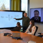 Local and remote members of the Microsoft Invoice Service team meet in a conference room to discuss the Modern Invoice API project. They are collaborating using a digital whiteboard and Microsoft Teams.