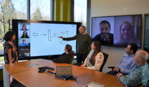 Local and remote members of the Microsoft Invoice Service team meet in a conference room to discuss the Modern Invoice API project. They are collaborating using a digital whiteboard and Microsoft Teams.