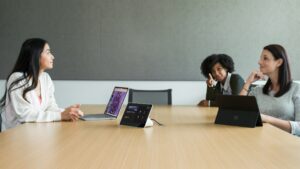 Three females in a medium conference room featuring a Poly Teams Meeting Rooms touch display with the Teams Meeting pre-join screen in view. Two Surface Devices in view.