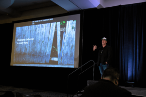 Ken Sexsmith stands on stage as he gives his Ignite 2019 presentation. The slide reads, “Changing behavior is really hard.”