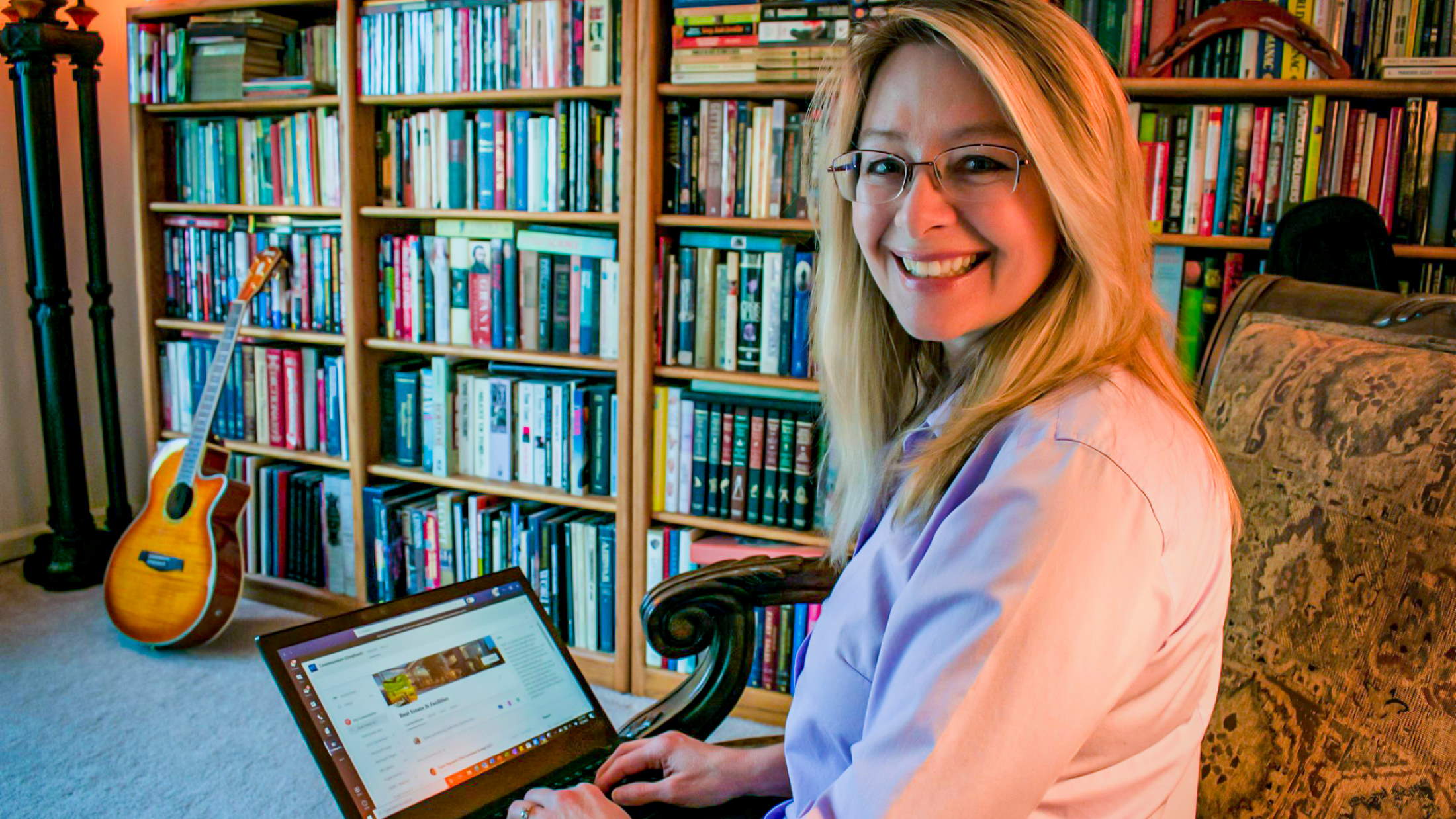 Gina Dyer sits in a library and smiles at the camera while holding her laptop.